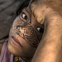 Em primeiro plano, desfocado, aparecem uma mão e o antebraço de um adulto que pinta o rosto de uma criança do povo Kaxinawá com grafismos tradicionais  na cor preta utilizando um palito. No plano principal aparece apenas o rosto da criança que olha para câmera.